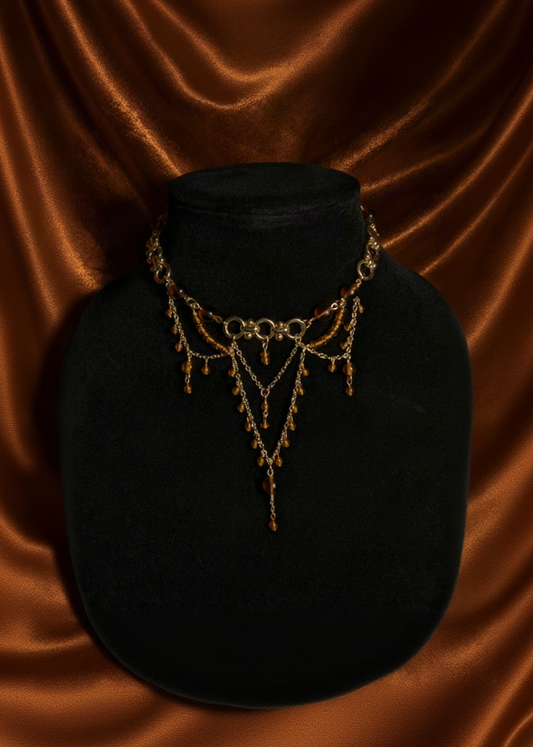 An ornate gold-toned chain necklace with dangling amber beads and a central heart-shaped charm, displayed on a black velvet bust against a draped brown velvet background.