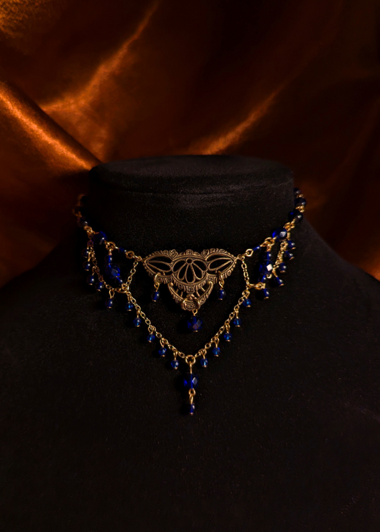 An ornate gothic-style choker necklace with a gold-toned filigree heart design and dangling dark blue beads, displayed on a black velvet bust against a draped brown satin background.