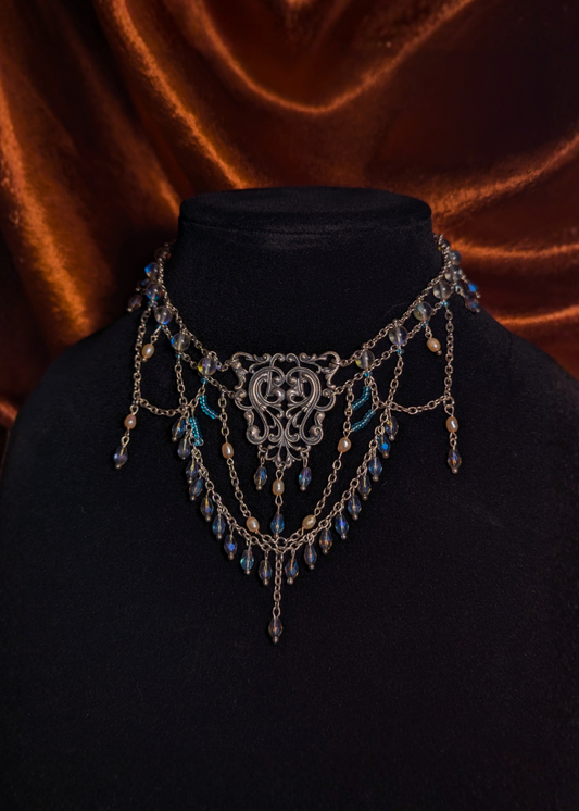 An ornate silver-toned filigree necklace with blue bead accents, displayed on a black velvet bust against a draped brown velvet background.