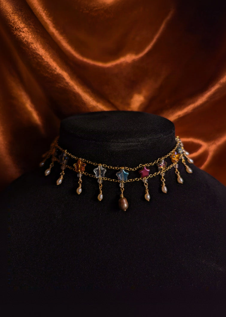 A gold-toned choker necklace with dangling charms, including star-shaped beads, teardrop beads, and small pearl-like drops, displayed on a black velvet mannequin bust against a draped copper-brown fabric background.