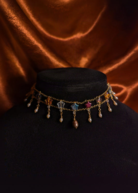 A gold-toned choker necklace with dangling charms, including star-shaped beads, teardrop beads, and small pearl-like drops, displayed on a black velvet mannequin bust against a draped copper-brown fabric background.